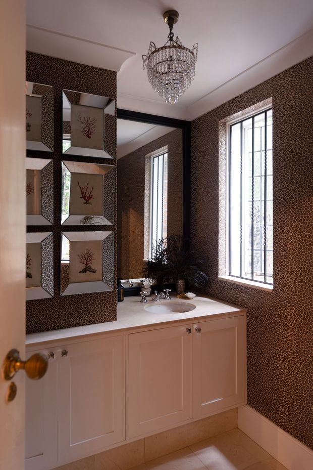 Elegant traditional bathroom viewed through an open door, featuring a white built-in vanity with pale stone benchtop and integrated basin, patterned brown wall covering, tall leaded-style windows, mirrored display niches, and an ornate crystal chandelier. The room has refined, decorative detailing and warm natural light.