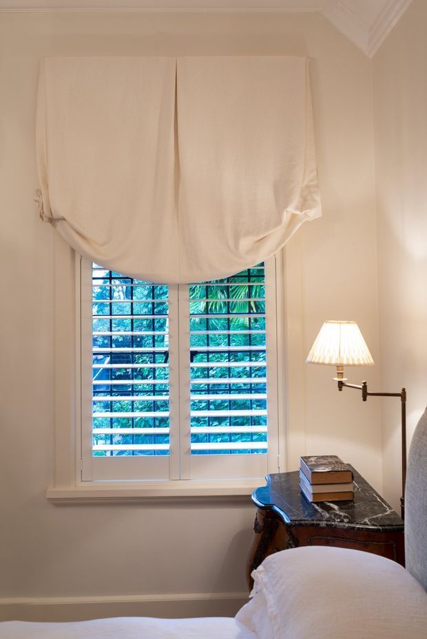 A softly lit bedroom corner featuring a tall louvered window with a loose cream Roman blind, pale walls and trim, a marble-topped dark wood bedside table, a traditional adjustable wall lamp, and part of a neatly made bed. Lush greenery is visible outside the window.