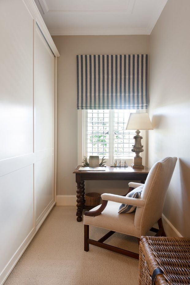 A narrow, refined home office or study with a dark wood writing desk positioned beneath a window. The room features warm beige walls, cream carpet, white panelled built-in cabinetry, plantation shutters, a striped Roman blind, an upholstered wooden armchair, and a traditional table lamp. White decorative ceiling mouldings and classic detailing add an elegant, tailored character.