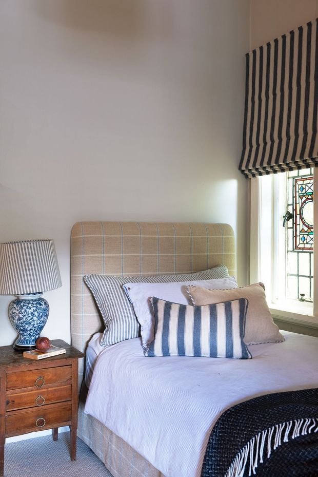 A cozy, traditionally styled bedroom featuring a single upholstered bed with a beige and pale blue checked headboard, layered blue-and-white striped bedding and cushions, and a dark woven throw at the foot. A wooden bedside table holds a blue-and-white ceramic lamp, while a striped Roman blind and decorative stained-glass window add character.