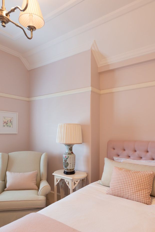 A softly styled bedroom with blush-pink walls, white decorative wall moulding and ornate ceiling cornices. The room contains a tufted upholstered bed, patterned cushions, a classic armchair, and a decorative side table with a ceramic lamp. The overall look is elegant, feminine, and traditionally detailed, with warm pastel tones and layered textiles.