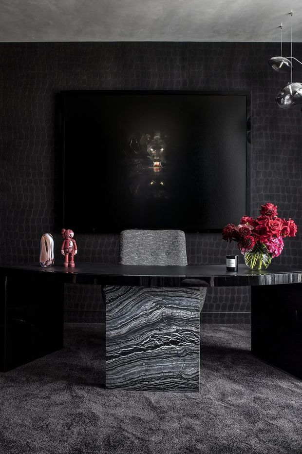A dramatic, dark home office or study featuring a long black built-in desk, a central sculptural stone pedestal or support with bold black-and-white veining, textured charcoal wall covering, dark carpet, and a large black artwork or screen. A grey upholstered chair sits at the desk, with sculptural pendant lights, small decorative objects, and a vivid red floral arrangement adding contrast.