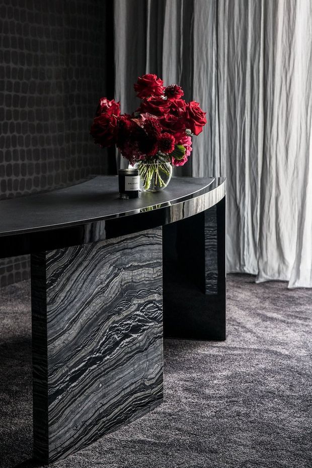 A dramatic, moody interior vignette featuring a sculptural black console table with a highly veined natural stone panel and glossy dark surface. A clear glass vase of deep red flowers sits beside a small dark container. Floor-to-ceiling sheer curtains, a dark patterned wall, and charcoal carpet create a luxurious monochromatic setting with rich red floral contrast.