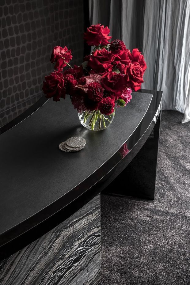 A moody, luxurious interior vignette featuring a black curved dining or console table with a dark textured top and reflective trim. A clear glass vase filled with deep red roses and dark flowers forms the focal point, set against a black patterned wall and sheer grey curtains. Dark carpet and a dramatic veined stone surface reinforce the high-contrast, monochromatic styling.