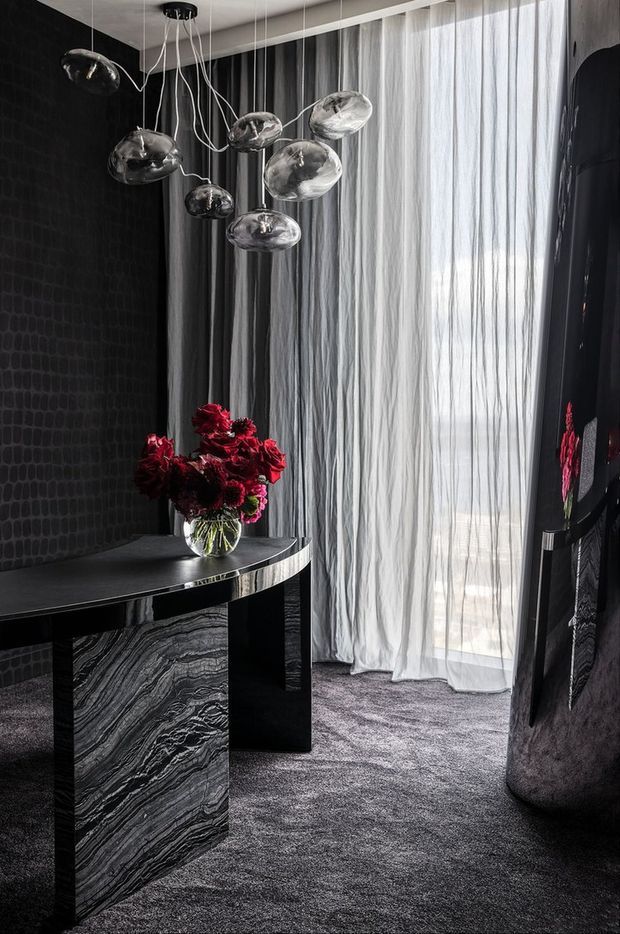 A dramatic, moody interior corner featuring a sculptural black stone console table with a boldly veined stone pedestal, dark textured wall covering, charcoal carpet, sheer floor-to-ceiling curtains, and a cluster of reflective sculptural pendant lights. A vase of deep red flowers provides the main colour accent, while a large dark reflective sculptural object stands at right.