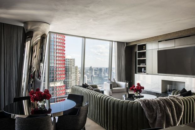A sophisticated high-rise living room and dining area with expansive floor-to-ceiling city glazing. The space features a large curved channel-tufted sectional sofa, a dark round dining table with upholstered chairs, a marble fireplace surround, dark built-in media cabinetry, full-height curtains, and sculptural reflective wall columns. Red floral arrangements provide vivid accents against a neutral grey, black, and white palette.