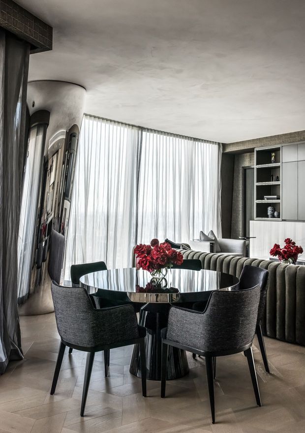 A sophisticated open-plan dining and living area with a round reflective dining table surrounded by dark upholstered chairs. A curved, channel-tufted banquette separates the dining space from a lounge area, while sheer floor-to-ceiling curtains soften the daylight from expansive glazing. The room features sculptural curved walls, dark built-in cabinetry, stone or plaster surfaces, and deep red floral arrangements as accents.