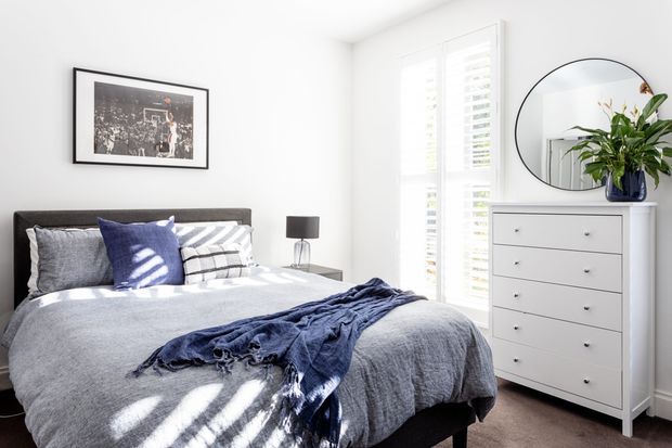 Bright, neatly styled bedroom with a charcoal upholstered bed, layered grey and navy bedding, white walls, a white chest of drawers, round wall mirror, bedside lamp, framed artwork, and plantation shutters. Strong natural light creates striped shadows across the bed.