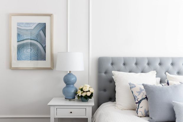 A softly styled bedroom vignette featuring a grey button-tufted upholstered bedhead, layered white and pale blue bedding, and a white bedside table with a blue ceramic lamp and flowers. The light grey wall is accented by framed blue-toned artwork and simple white trim.
