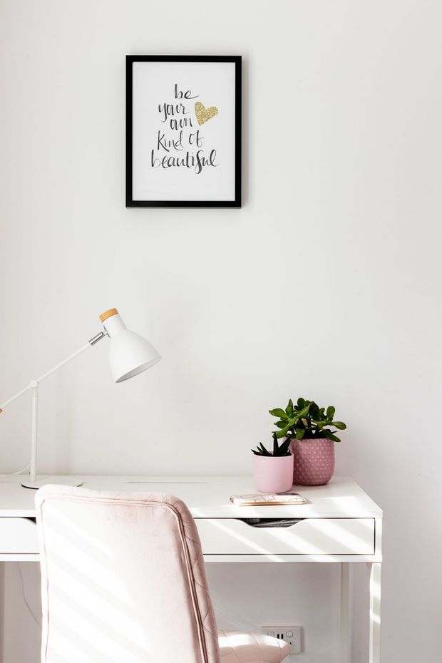 Bright minimalist home workspace featuring a white desk with drawers, white angled desk lamp, blush pink upholstered chair, and two potted plants. A black-framed inspirational print hangs on the white wall above the desk, with soft natural light creating diagonal shadows.