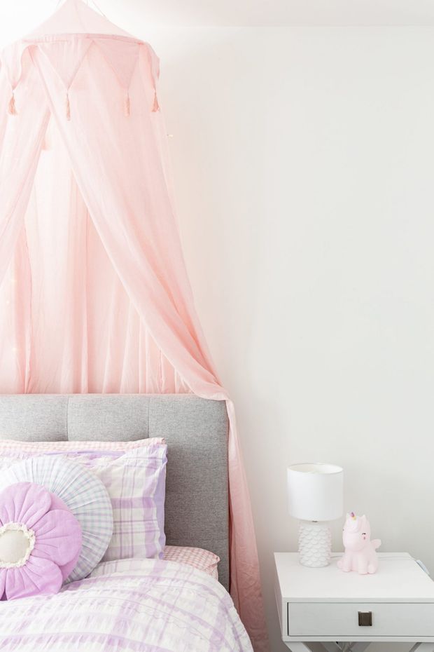 Bright, softly styled child's bedroom featuring a grey upholstered bedhead, pastel lilac and pink patterned bedding, and a sheer blush-pink canopy draped above the bed. A white bedside table holds a small white lamp and pink decorative figurine. The room has pale walls and a light, playful aesthetic.