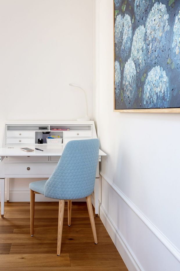 A compact, bright home office corner with a white built-in desk and drawer storage, a pale blue upholstered mid-century-style chair, warm timber flooring, white walls with decorative wainscoting, and a large blue floral artwork in a timber frame.