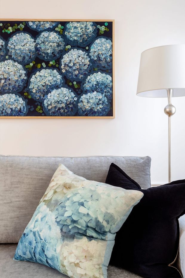 Close-up interior detail of a contemporary living room, featuring a light grey upholstered sofa with patterned blue floral cushions and a black cushion. A large framed blue floral artwork hangs on the pale wall above, beside a slim metallic floor lamp with a white drum shade. The styling is calm and refined with a soft blue, grey, white, and black palette.