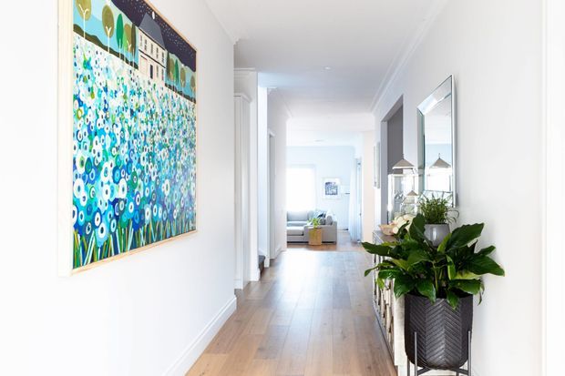 A bright, long residential hallway with white walls, decorative ceiling cornices, and warm timber flooring. A large colourful floral artwork hangs on the left wall, while a tall planter and large mirror decorate the right side. The corridor leads toward a softly furnished living area at the far end.