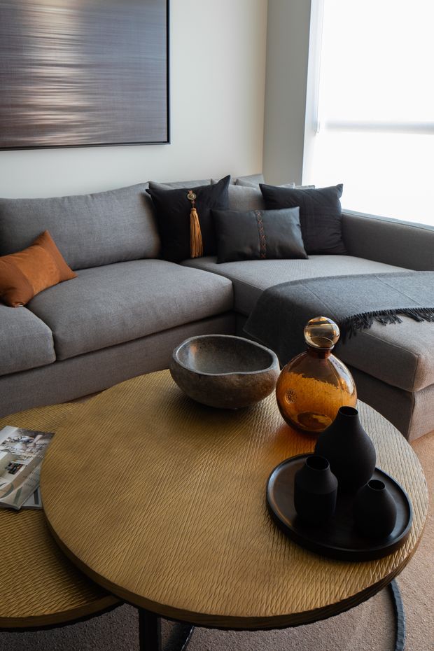 A close interior view of a refined living room featuring a large grey L-shaped sectional sofa with dark and rust-toned cushions, a textured round wood coffee table, sculptural black ceramic vessels, an amber glass vase, and a dark grey throw. A contemporary abstract artwork hangs on the light neutral wall beside a bright window with a roller blind. The room has a warm, layered, sophisticated atmosphere.