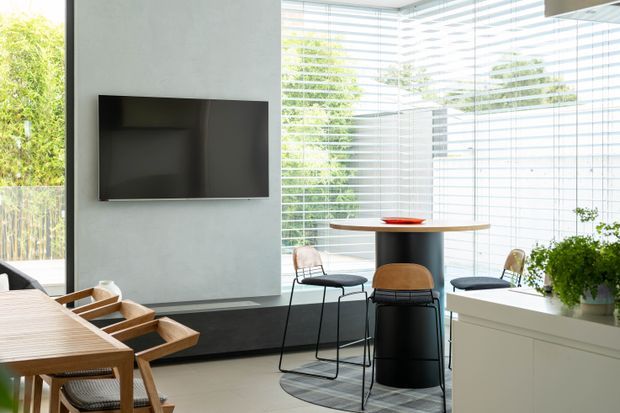 Bright contemporary open-plan interior with a compact dining and bar area beside floor-to-ceiling glazed walls fitted with horizontal blinds. A wall-mounted television sits on a pale textured wall, while a timber dining table, black cylindrical island or bar table, wire-frame stools, and built-in white cabinetry define the space. Greenery is visible outside and in a planter, creating an airy indoor-outdoor atmosphere.