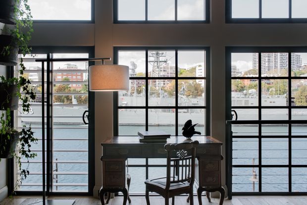 A refined interior workspace or console area positioned in front of expansive black-framed industrial-style glazing. A traditional dark wood desk with carved detailing and a matching chair sits beneath a large pendant lamp, while a potted climbing plant softens the space. Through the windows, a waterfront promenade, water, trees, and urban buildings are visible. The room has a high ceiling and strong indoor-outdoor connection.