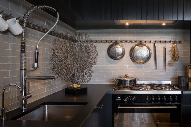 A dark, characterful kitchen with charcoal cabinetry, black benchtops, grey subway-tile walls, and a large stainless-steel range cooker. A deep undermount sink sits beneath a spring-coil faucet, while copper pans and utensils hang from rails against the tiled backsplash. Decorative branch-like wall art, pendant crockery, and warm recessed lighting add rustic industrial character.