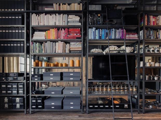 A dark, industrial-style built-in shelving and storage wall filled with books, binders, labelled archival boxes, records, display objects, ceramics, glassware, and organised storage containers. A black ladder leans against the central shelving, with a television or dark display panel integrated into the lower middle section. The space has black metal framing, dark timber shelves, and a warm timber floor, creating a highly organised library, studio, or home office storage area.