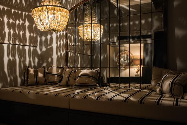A moody, luxurious bedroom with a wide built-in upholstered platform bed extending across the foreground. The bed is dressed in warm taupe and beige striped bedding with coordinated cushions. Dark gridded glass-panelled doors or room dividers form a reflective backdrop, while ornate crystal pendant lights cast dramatic patterned shadows across the walls and ceiling. The palette is warm and dark, with rich reflective detailing and an intimate atmosphere.