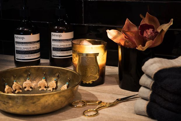 Close-up still life of a warmly lit bathroom vanity or benchtop, featuring Aesop bottles, a brass bowl with small decorative objects, a glowing candle, a dark vase with dried flowers, folded towels, and grooming scissors. The scene has a refined, spa-like atmosphere with a dark tiled backsplash and warm stone or concrete-look surface.