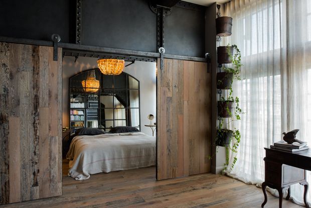 Industrial-style bedroom with rustic timber sliding barn doors, exposed dark metal framing, and a large black-framed arched mirror reflecting the bed. Warm pendant lighting, timber flooring, sheer full-height curtains, and a tall plant display create a moody yet inviting interior.