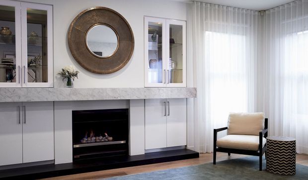 Refined living room featuring a white built-in media and storage wall with glass-front display cabinets, a marble-look horizontal shelf, a black fireplace insert, and a large round metallic-framed mirror. Sheer floor-to-ceiling curtains provide soft natural light, while a cream upholstered armchair and patterned cylindrical side table complete the seating area.