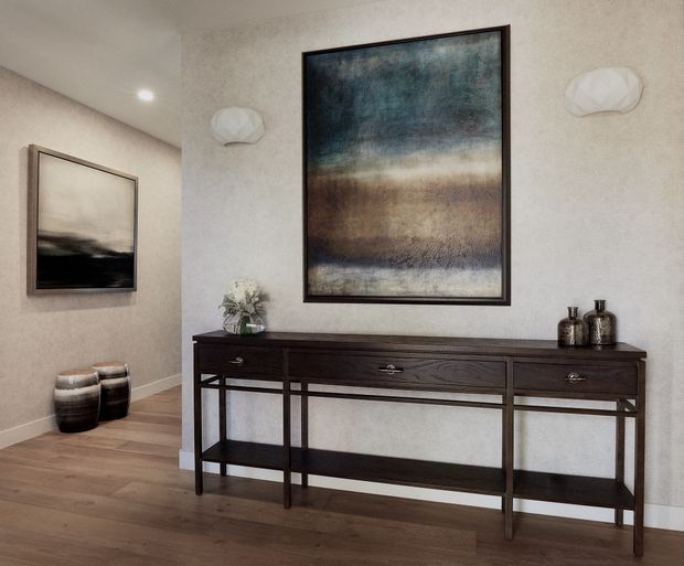 A refined hallway or entryway featuring a long dark timber console table with open lower shelving, abstract artwork, sculptural wall lights, decorative vessels, and warm timber flooring. The neutral textured walls and restrained furnishings create a calm, contemporary atmosphere.