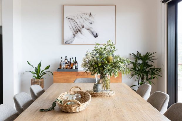Bright contemporary dining room with a large light timber dining table surrounded by upholstered chairs. A low timber sideboard with bottles sits against a white wall beneath framed horse artwork, while abundant leafy floral arrangements and indoor plants add a natural accent. Full-height glazed doors or windows are visible to the right.
