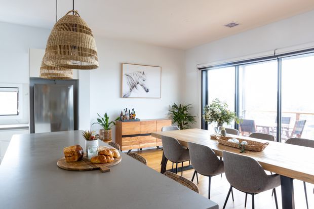 Bright open-plan kitchen and dining area with a large pale island bench in the foreground, stainless-steel refrigerator and white cabinetry at left, a timber sideboard, indoor plants, and a timber dining table with upholstered chairs beside expansive glazing. A woven pendant light and natural timber furnishings add a relaxed contemporary feel, with an outdoor terrace visible beyond the windows.