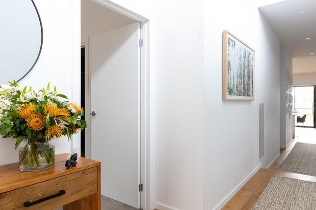 Bright, minimalist residential hallway with white walls, timber flooring, and a narrow console table topped with a glass vase of flowers. An open doorway leads to an adjacent room, while framed artwork and a long runner define the corridor.