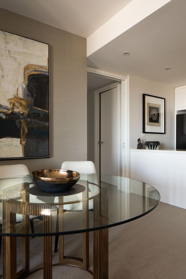 A refined dining area with a round glass-top dining table, sculptural bronze-toned bowl, upholstered white dining chairs, and a large abstract artwork. The room has textured taupe wall covering, white trim and built-in cabinetry, neutral carpeting, recessed lighting, and a partially visible adjoining hallway or room.