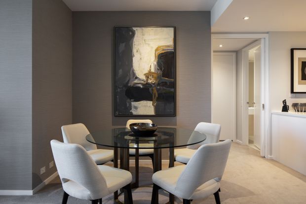 A refined dining area with a round glass dining table and four upholstered white dining chairs. The room features textured dark grey wallcovering, a large abstract artwork, dark timber or metal table framing, pale carpet, recessed lighting, and a bright white built-in cabinet area leading toward a hallway and adjacent spaces.