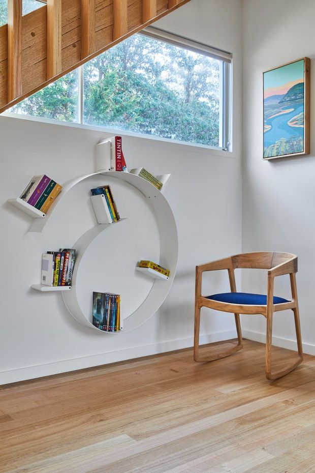 A bright, compact reading nook or home office corner with white walls, warm timber flooring, and a distinctive circular wall-mounted bookshelf filled with books. A sculptural timber chair with a blue upholstered seat sits beside the shelving. The room features a large angled window with views of leafy trees, exposed warm timber ceiling or roof beams, and a colourful framed landscape artwork. The overall design is playful, refined, and mid-century influenced.