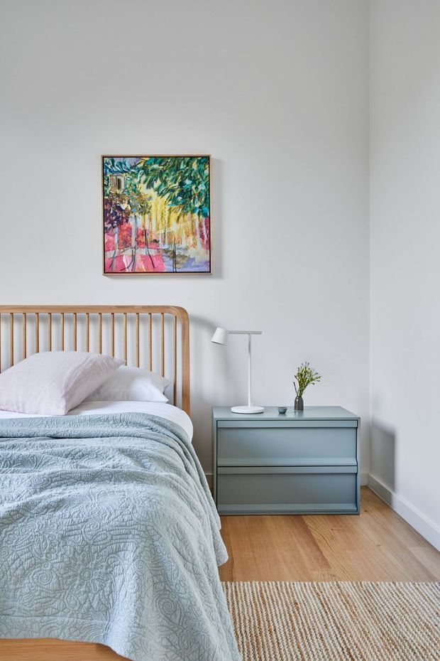 A bright, calm bedroom with a timber slatted headboard, pale blue textured bedding, a blue-grey bedside chest, and a slim white floor lamp. The room has white walls, warm timber flooring, a woven neutral rug, and colourful framed artwork above the bed.
