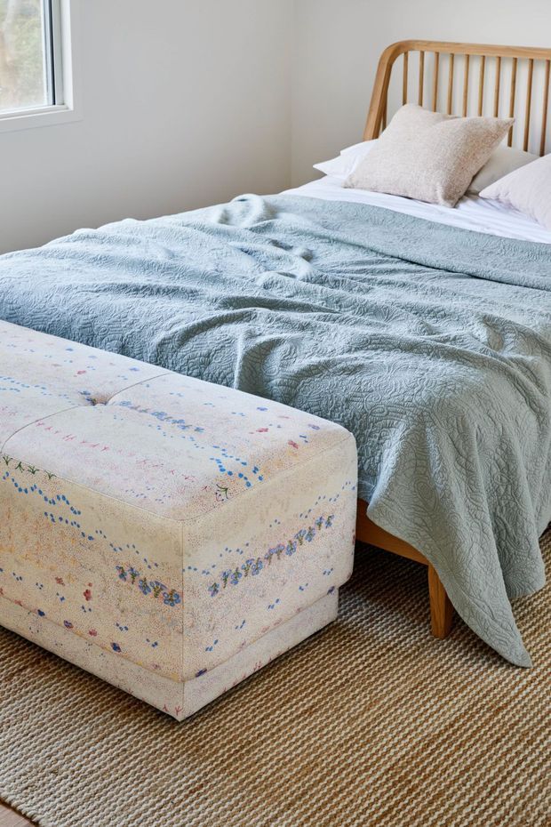 A bright, softly styled bedroom featuring a timber spindle-headboard bed dressed in pale blue textured bedding and light pillows. A large upholstered ottoman with a delicate multicolour floral pattern sits at the foot of the bed, over a natural woven carpet. White walls and a simple white-framed window create an airy, relaxed atmosphere.