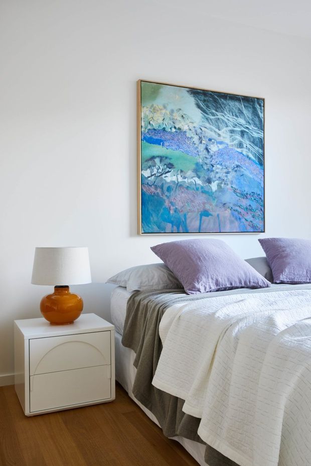 Bright, softly styled bedroom featuring a neatly made bed with layered white and grey bedding, lavender pillows, a white curved-front bedside table, and an amber glass table lamp. A large blue and lavender abstract artwork hangs above the bed. The room has plain white walls and warm timber flooring.