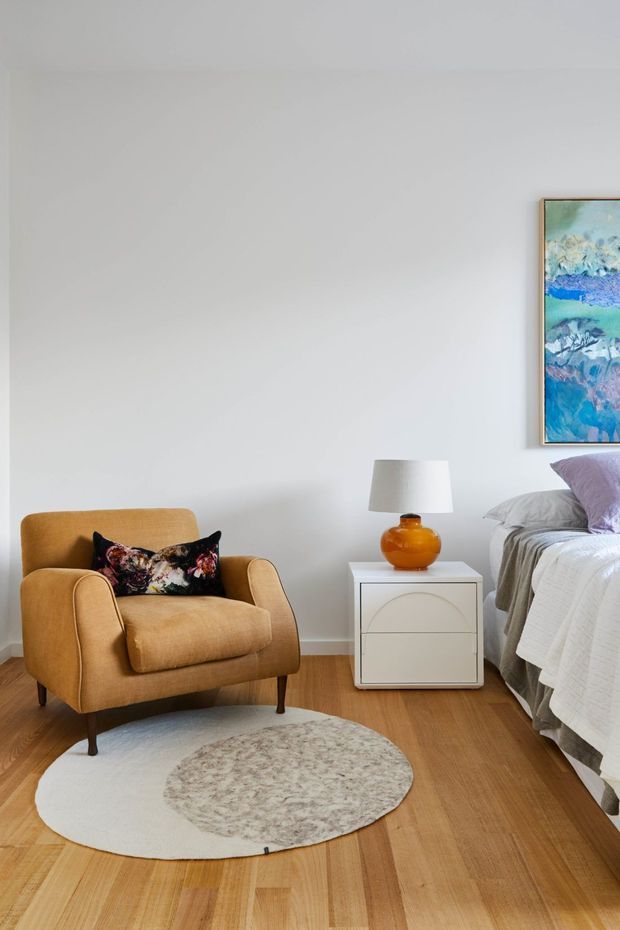 A softly styled bedroom corner with a warm mustard upholstered armchair, patterned cushion, white bedside table, amber glass lamp, and partially visible bed. Light timber flooring, pale walls, a round layered rug, and colourful framed artwork create a calm, contemporary interior.
