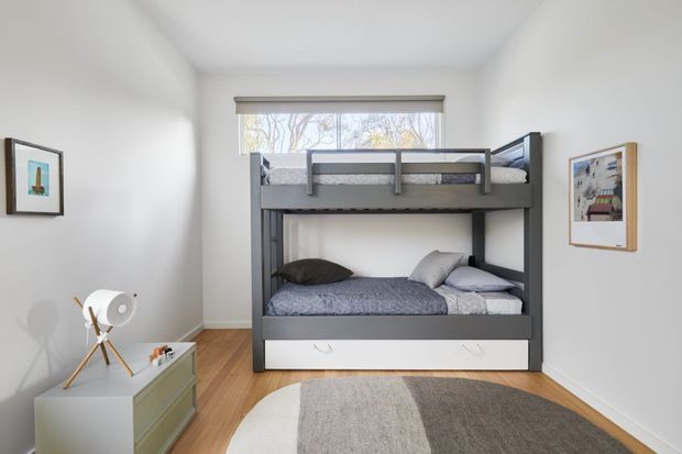 Bright, compact bedroom featuring a grey built-in bunk bed with a lower double bed and upper bunk, white integrated drawers, pale walls, timber flooring, a low light-coloured storage unit, framed artwork, and a soft neutral rug. Daylight enters through the rear window, creating a clean and practical children's or guest bedroom setting.