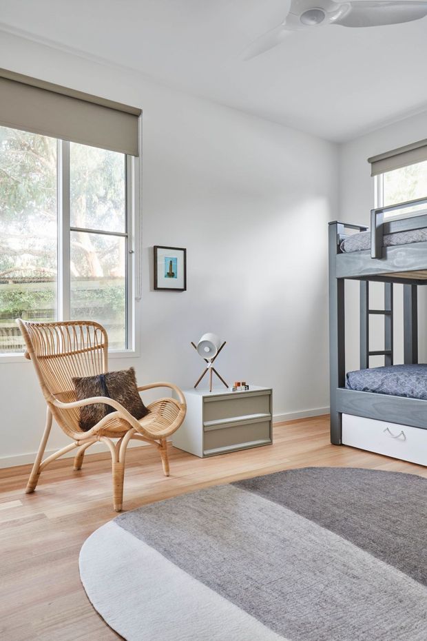 A bright, tidy children's bedroom with white walls, pale timber flooring, a large window with roller blinds, a grey-and-black bunk bed, woven rattan lounge chair, compact bedside cabinet, small framed artwork, and a large grey and off-white area rug. A ceiling fan is visible overhead.