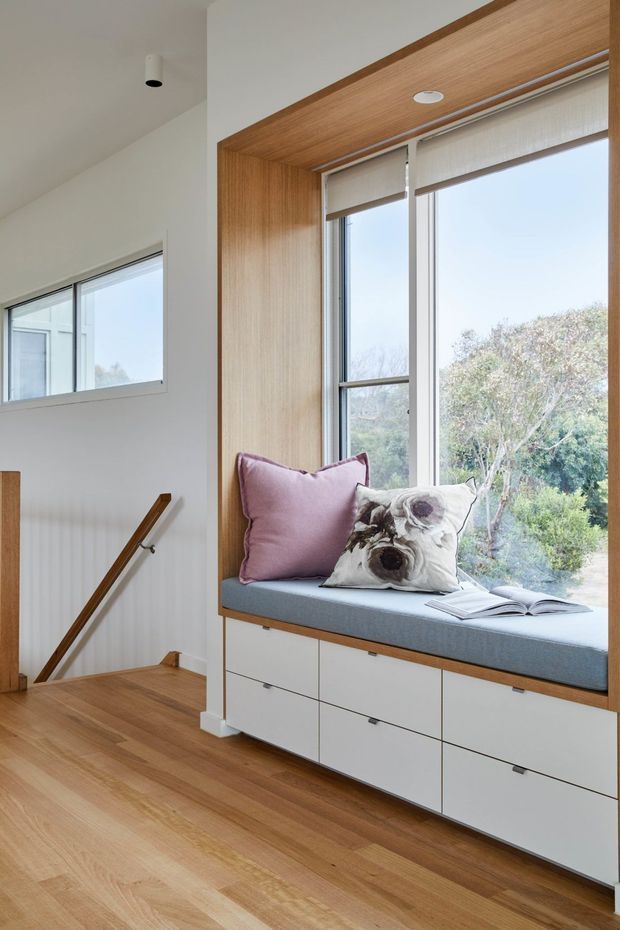 Bright interior landing beside a stairway, featuring a large timber-framed window seat with built-in white drawers, blue-grey upholstered cushion, decorative pillows and an open book. Natural timber flooring, white walls, slim window frames and a timber handrail create a clean, warm contemporary feel.