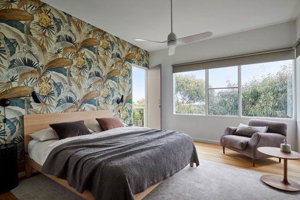 A bright, contemporary bedroom with a large bed positioned against a tropical botanical feature wall. The room has light timber flooring, neutral bedding, a large window with a roller blind, a ceiling fan, and a mauve upholstered lounge chair with a small round side table. Natural light and garden views create an airy atmosphere.