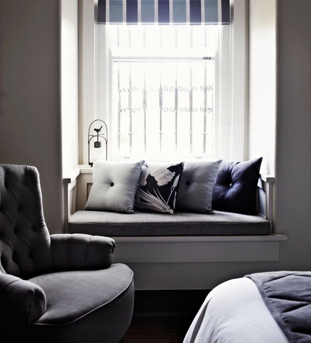 A cozy bedroom corner featuring a built-in window seat beneath a bright window with decorative security bars and a navy-and-white striped Roman blind. The seat has grey upholstery and a mix of grey, black, and white cushions, accompanied by a large tufted grey armchair in the foreground. Dark timber flooring and muted grey walls create a calm, refined atmosphere.