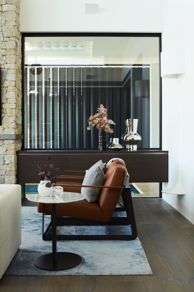 A refined living room vignette featuring a cognac leather lounge chair with dark timber or black framing, positioned on a muted blue-grey rug beside a small round side table. A dark timber console sits beneath a large black-framed interior window or mirror, with sculptural glassware and dried floral decor. The room includes a prominent rough natural-stone wall, white plaster walls, dark timber flooring, and a streamlined white sculptural fireplace or built-in feature.