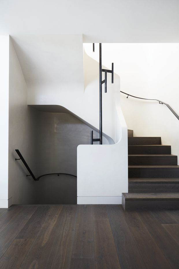 A minimalist interior stairway with dark timber flooring and matching dark timber treads, white sculptural curved balustrade walls, and slim black metal handrails. The view looks across a split-level stair landing, with strong geometric lines, abundant white surfaces, and soft natural light.