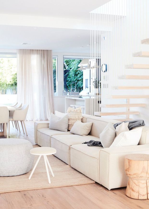 Bright, airy open-plan living area with a large cream sectional sofa, pale upholstered ottoman, light timber flooring, neutral rug, and a small white side table. A dining area and white kitchen are visible behind the seating, with abundant glazing, sheer curtains, greenery outside, and a floating timber stair screened by vertical white rods.