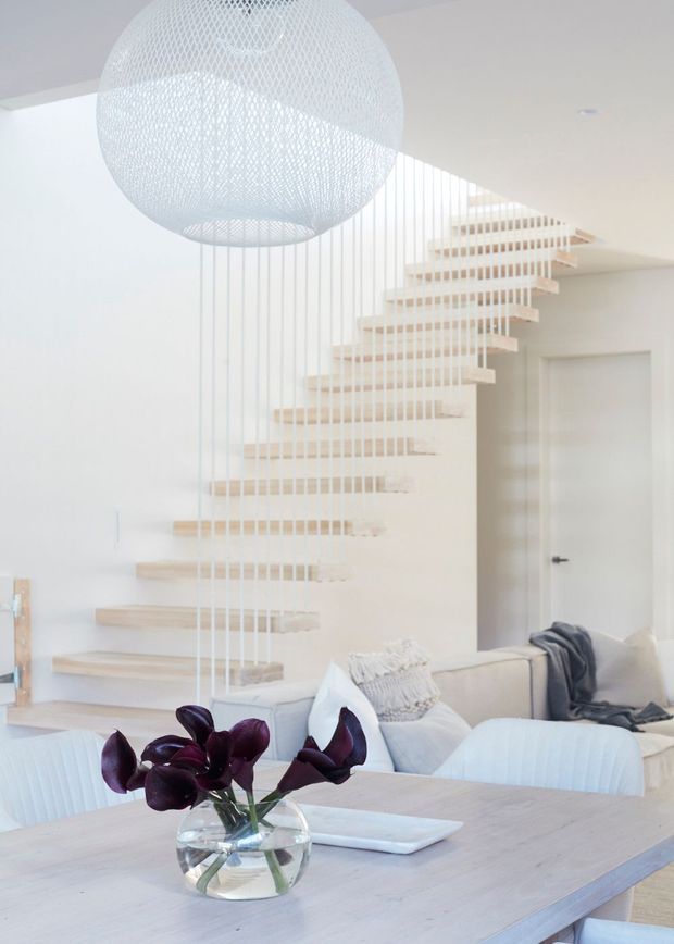 Bright, minimalist open-plan living and dining area featuring a floating timber staircase with white vertical balustrade cables, white walls, pale timber finishes, a large woven spherical pendant light, and soft neutral seating around a pale dining table. Dark purple flowers provide a small accent in the foreground.
