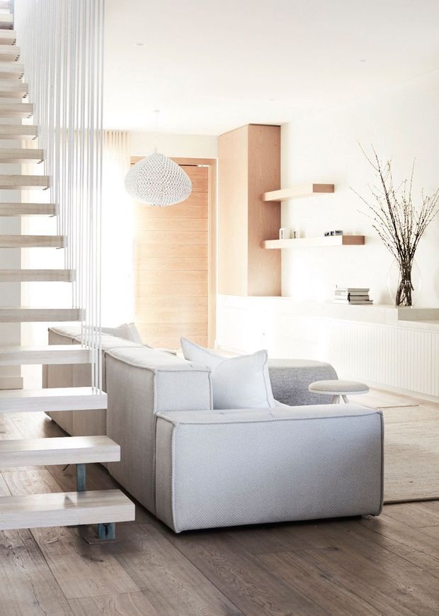 Bright, minimalist living room with a pale grey sofa, light timber flooring, white walls, and a floating staircase with slender vertical balustrade rods. Built-in white ribbed cabinetry runs along the rear wall, beside a warm timber pivot door and a tall timber feature cabinet with simple floating shelves. A woven pendant light, small round side table, books, and decorative branches add restrained detail.