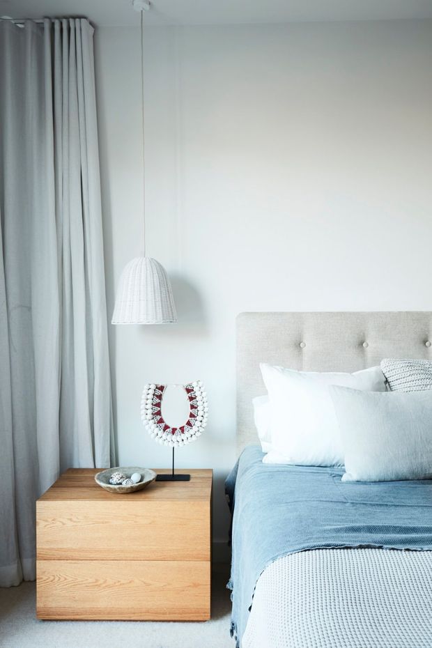 A softly lit, calm bedroom corner featuring a pale upholstered button-tufted headboard, white pillows, a light blue bedcover, floor-length light curtains, and a simple woven pendant light. A light timber two-drawer bedside table holds a small decorative object and a beaded ornament.