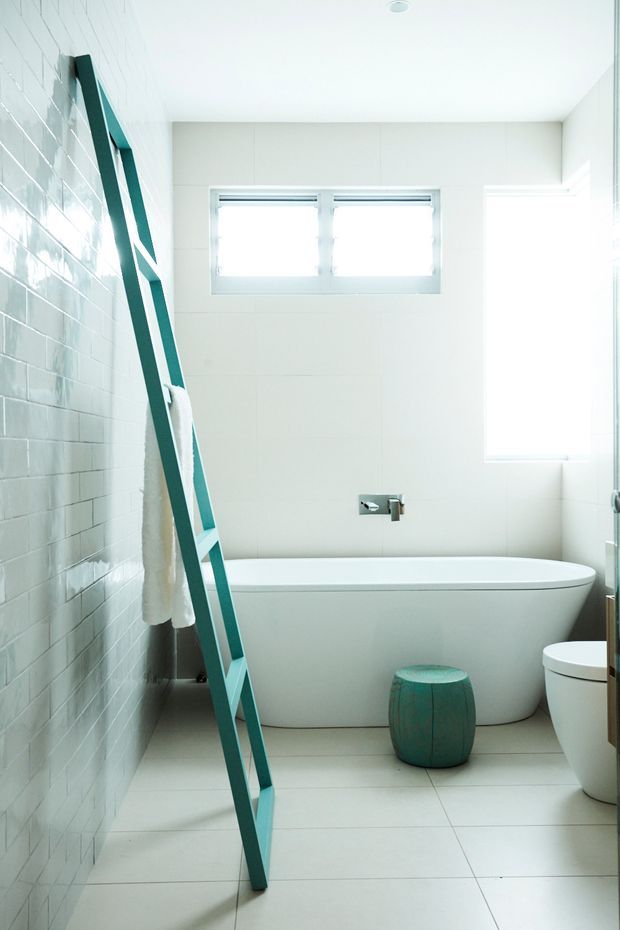 Bright, compact contemporary bathroom with a freestanding white bathtub, white tiled walls and floor, high horizontal windows, a white toilet, and a distinctive teal leaning towel ladder with a matching teal stool. The space is clean, light-filled, and minimally styled.